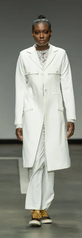 Model wearing a white coat and pants with brown sneakers on a runway.