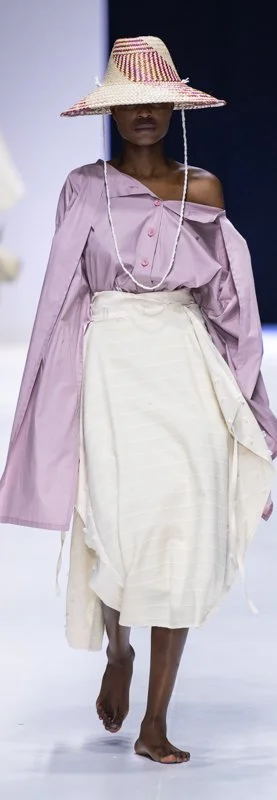 Model on runway wearing oversized purple shirt, white skirt, broad-brimmed straw hat, and necklace.