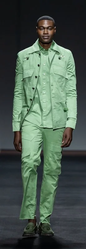Model wearing a light green outfit with tailored jacket, shirt, and pants on a runway.