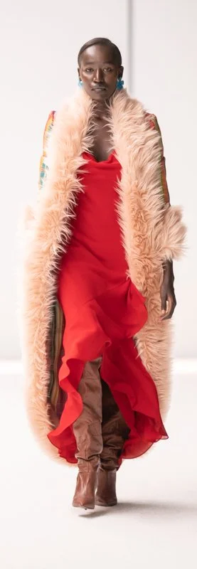 Model wearing a red dress with a long, fluffy coat and brown boots on a runway.