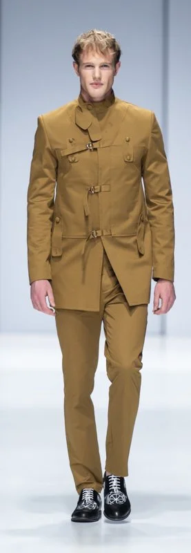 Model in brown tailored suit with high collar and toggle details, wearing black shoes on a runway.