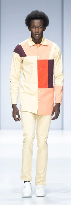 Model walking on runway wearing a color-blocked shirt and light-colored pants in a fashion show.