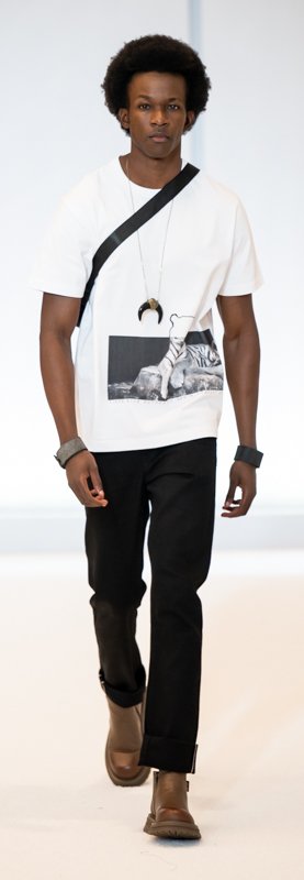 Fashion model wearing a white graphic t-shirt with black trousers and brown shoes, accessorized with a pendant necklace and wristbands, walking on a runway.