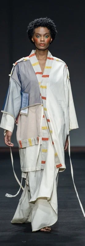 Model walking on runway wearing a multi-layered, geometric-patterned kimono-style outfit with a long, flowing design in neutral tones with subtle colorful accents.