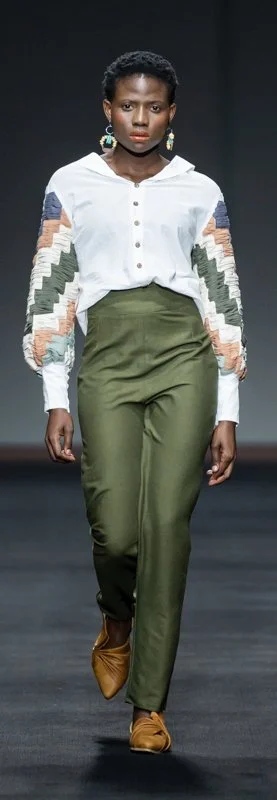 Model on runway wearing a white blouse with colorful zigzag sleeves, olive green high-waisted pants, and brown shoes.