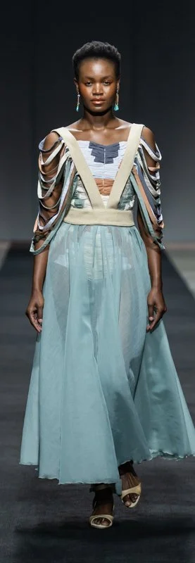 Model wearing a long, flowing pastel blue dress with cut-out shoulder designs, walking on a fashion runway.