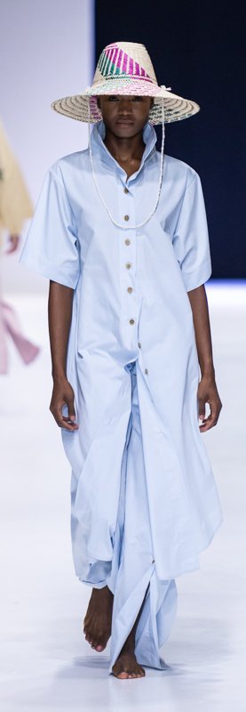 Model wearing long light blue button-up dress and wide-brimmed straw hat on runway