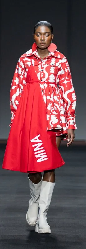 Model wearing a red and white patterned dress with "MINNA" text, paired with white boots on a runway.