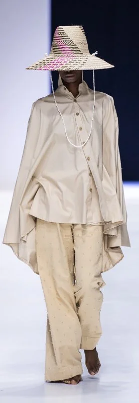 Model wearing an oversized beige shirt, wide-legged pants, and a large woven hat at a fashion show.