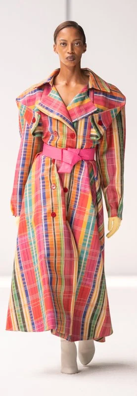 Person wearing a colorful plaid trench coat with pink belt on a runway.