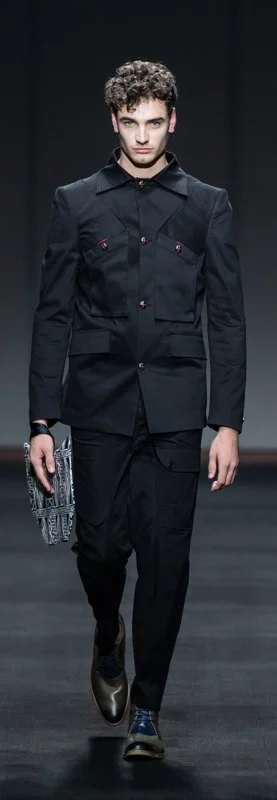 Model in a black tailored suit with a unique bag walking on a runway.