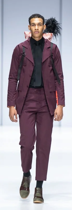 Model on runway wearing a burgundy suit and black shirt with feather detail, fashion show.