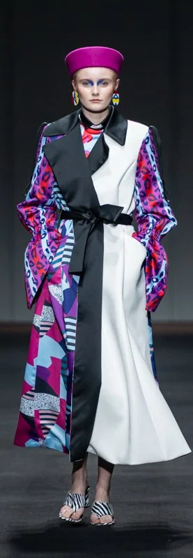Fashion model wearing a colorful mixed-pattern coat with black sash, vibrant pink hat, and bold geometric and animal prints walking on a runway.