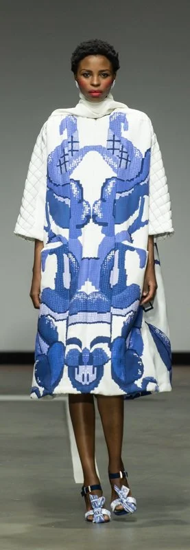 Model wearing a white and blue patterned dress with white quilted sleeves on a runway.