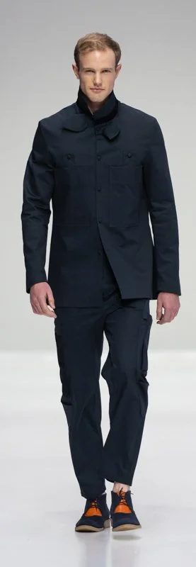 Model wearing a dark suit with unique pocket details, black shirt, and colorful sneakers on a runway.