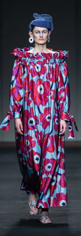 Model wearing a long floral dress with ruffled neckline on a runway.