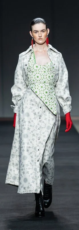 Model wearing a gray floral-patterned trench coat, green floral corset top, red gloves, red earrings, and black boots on a runway.