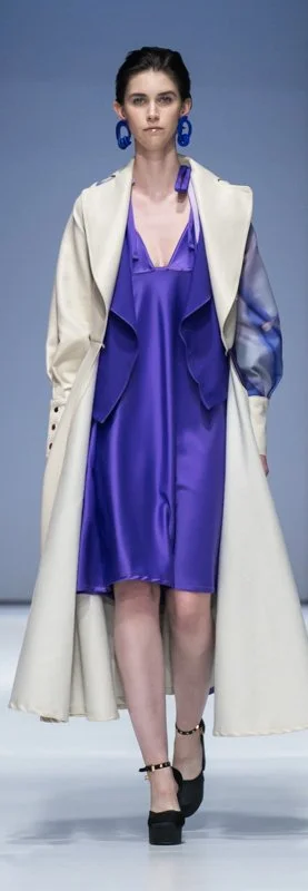 Model walking on a runway wearing a cream long coat over a purple dress, with blue earrings and black shoes.