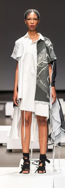 Fashion model on a runway wearing an asymmetrical, multi-layered white and gray dress with black lace-up sandals.