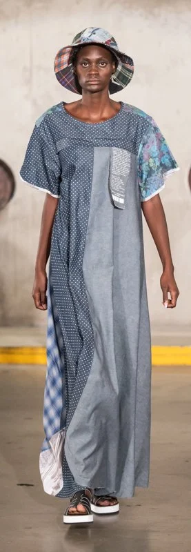 Model wearing a long patchwork dress and a plaid bucket hat on a runway.