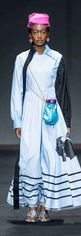 Model wearing a long-sleeved light blue dress with black accents and stripes, accessorized with a metallic blue bag, pink hat, and black and white striped shoes, walking on a runway.