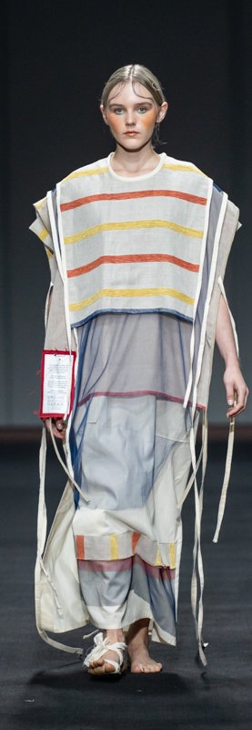 Model on runway wearing layered, striped tunic with loose ribbons