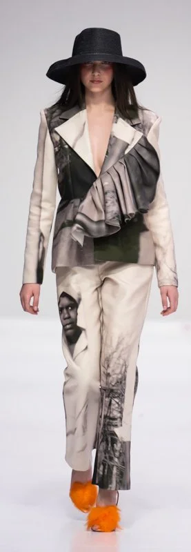 Model wearing a printed suit with a large black hat and orange fluffy shoes on a runway.