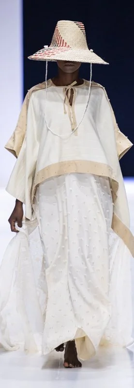 A model wearing a wide-brimmed straw hat and a flowing white outfit on a runway.