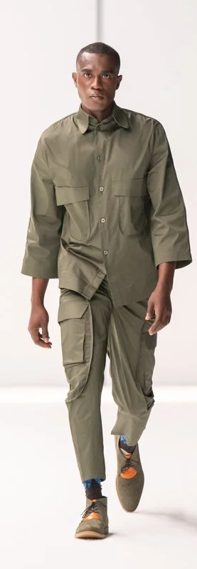 Model walking down the runway wearing an olive green outfit with oversized pockets, featuring a button-up shirt and cargo pants, paired with brown casual shoes.
