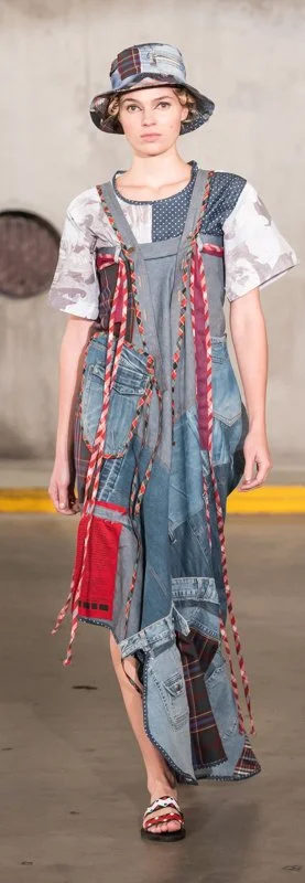 Model wearing a patchwork denim dress with mixed fabric patterns, including plaid and solid colors, with multiple pockets and red braided straps. The outfit is paired with a matching bucket hat and sandals, designed for a casual, eclectic fashion loo