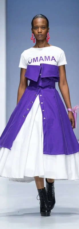 Model on runway wearing a white t-shirt with 'UMAMA' text and a purple layered garment over a white skirt, paired with black boots. The model also wears red earrings and carries a small accessory.