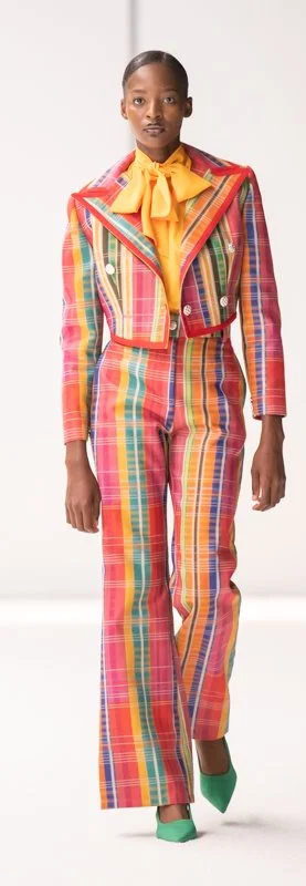 Model wearing a colorful plaid suit with a vibrant yellow blouse and green shoes on fashion runway.