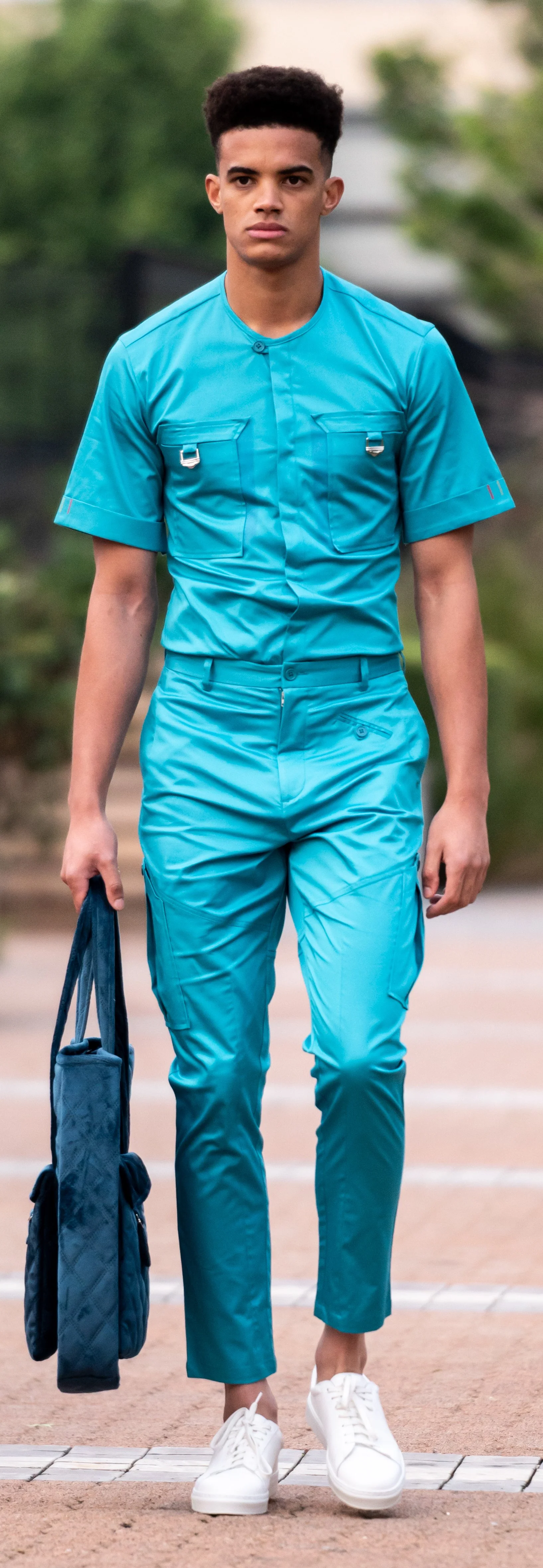 Model wearing a teal matching shirt and pants, holding a quilted blue bag, walking in white sneakers.