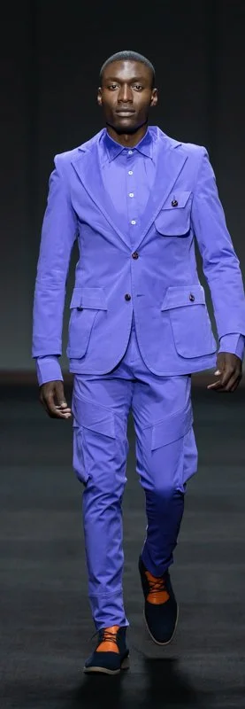 Model on runway wearing a bright blue suit with matching pants and a blue dress shirt, paired with black shoes and orange socks.