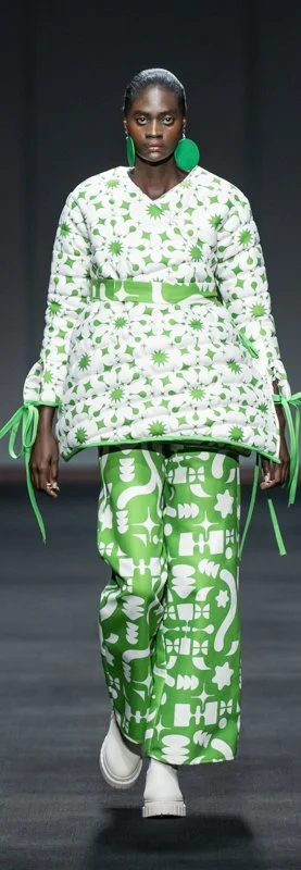 Model wearing a green and white patterned outfit on a runway, including a padded tunic with side ties and matching wide-leg pants, accessorized with large green earrings and white boots.