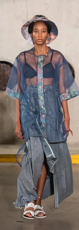 Fashion model wearing a sheer blue jacket with floral trim, denim skirt, and a plaid bucket hat on a runway.