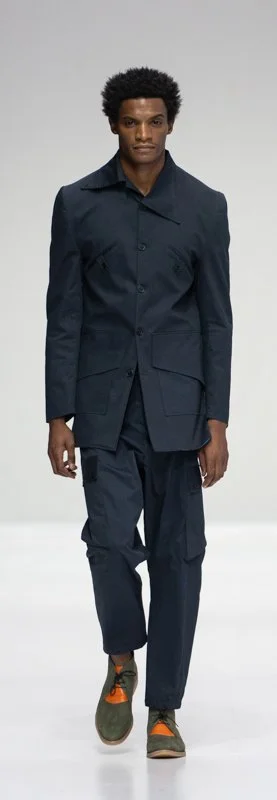 Model wearing a dark navy suit with matching pants and colorful shoes on a runway.