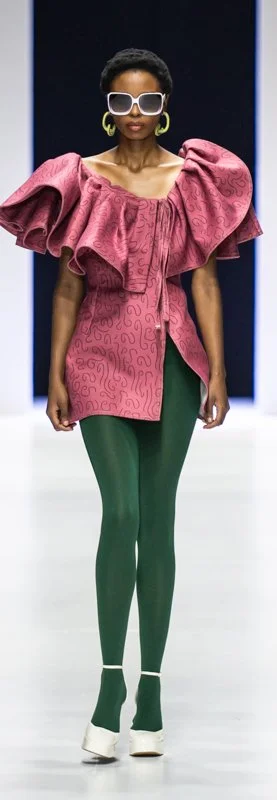 Fashion model on runway wearing pink ruffled top, green leggings, white high heels, large sunglasses, and yellow earrings.