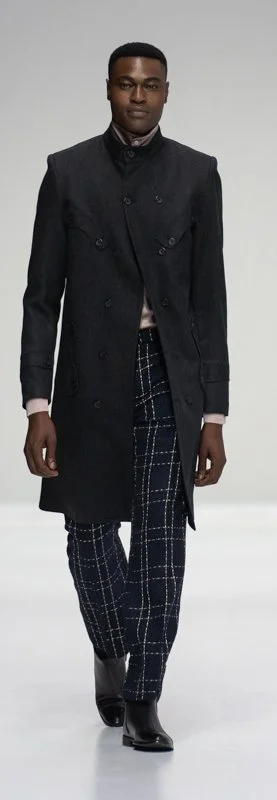 Man wearing a long black coat and plaid pants on a runway.