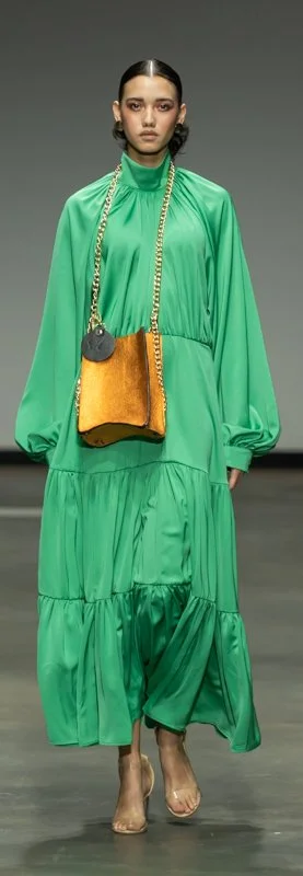 Model walking in fashion show wearing green long-sleeve dress, carrying orange handbag with gold chain.
