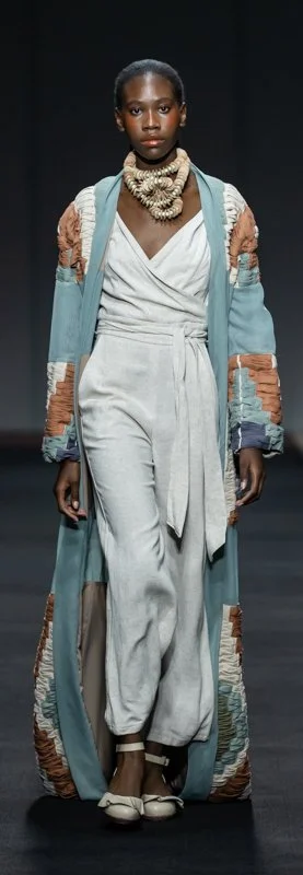 Model wearing a stylish long coat with textured patterns over a white wrap outfit, accessorized with a statement necklace and heels, walking on a runway.