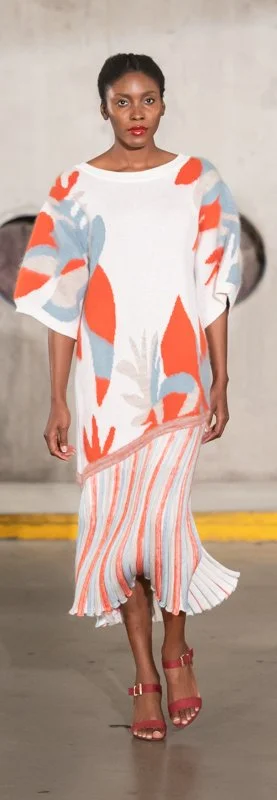 Model walking on runway in colorful knit outfit, featuring a white, orange, and blue abstract pattern top and matching striped skirt, with red sandals.