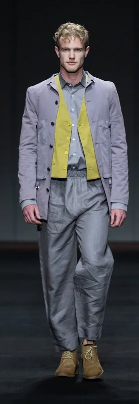 Model wearing purple jacket, yellow vest, gray shirt, baggy gray pants, and brown shoes on runway