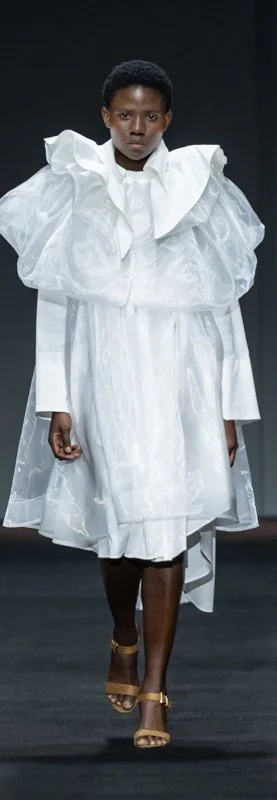 Model walking runway in avant-garde white dress with ruffled collar and transparent overlay.