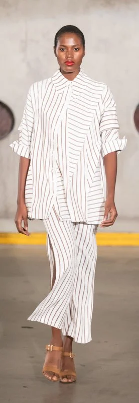 Fashion model in flowing white outfit with brown stripes and brown sandals walking on a runway.