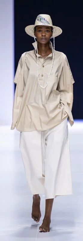 Model on runway wearing beige oversized shirt, white wide-leg pants, straw hat, and necklace