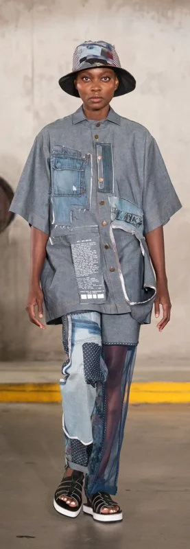 Model wearing oversized denim outfit with patchwork design, bucket hat, and sandals on runway.