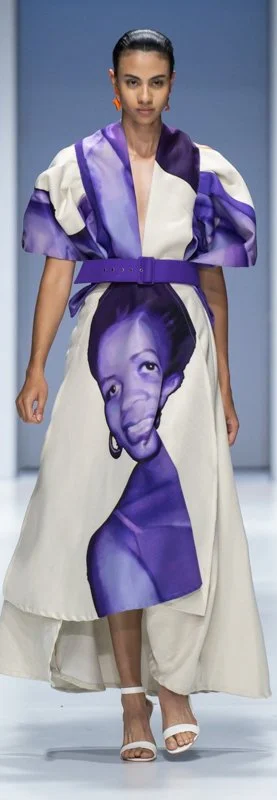 Model wearing a dress with a purple and white design, featuring a large portrait of a person on the skirt, walking on a fashion runway.