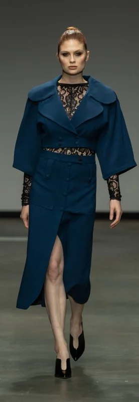 Model walking the runway in a blue coat with a high slit and black lace sleeves at a fashion show.