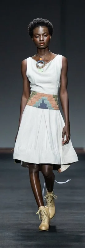 Model walking on runway wearing a sleeveless beige dress with geometric pattern in the waist and tan shoes.